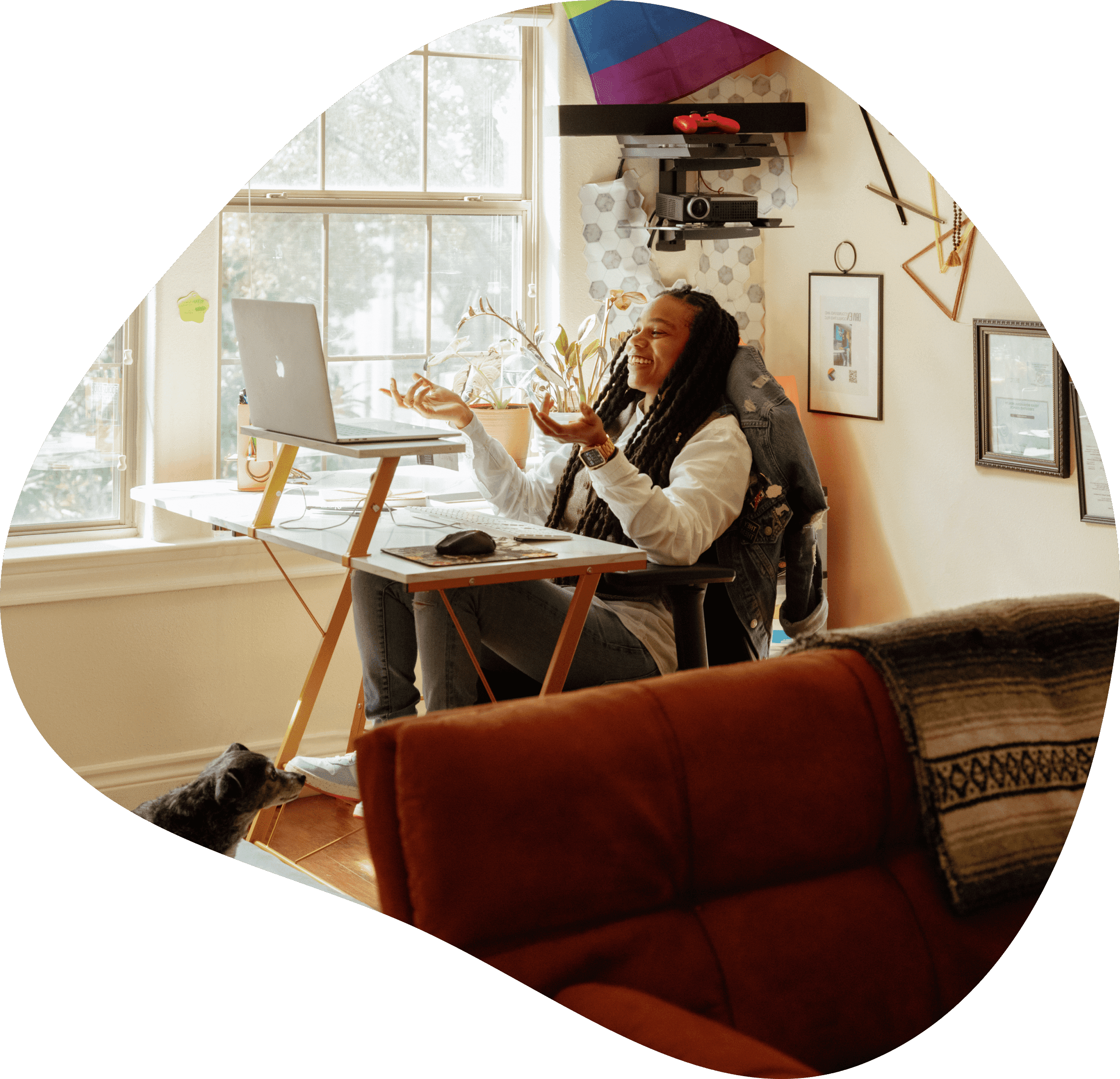 Black nonbinary mental health counselor smiling at their computer while sitting at their desk in a sunlit room with a dog at their feet.